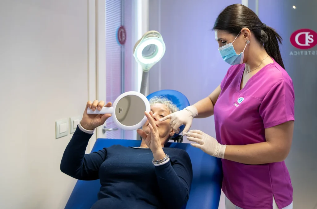 Paziente con capelli grigi ricci, seduta su una poltrona blu, si guarda allo specchio rotondo durante un consulto estetico. Accanto a lei, una professionista dello studio Dentas con camice rosa e mascherina le indica un punto del viso con guanti bianchi. Sullo sfondo, una lampada ad anello e il logo Dentas Estetica sulla vetrata.