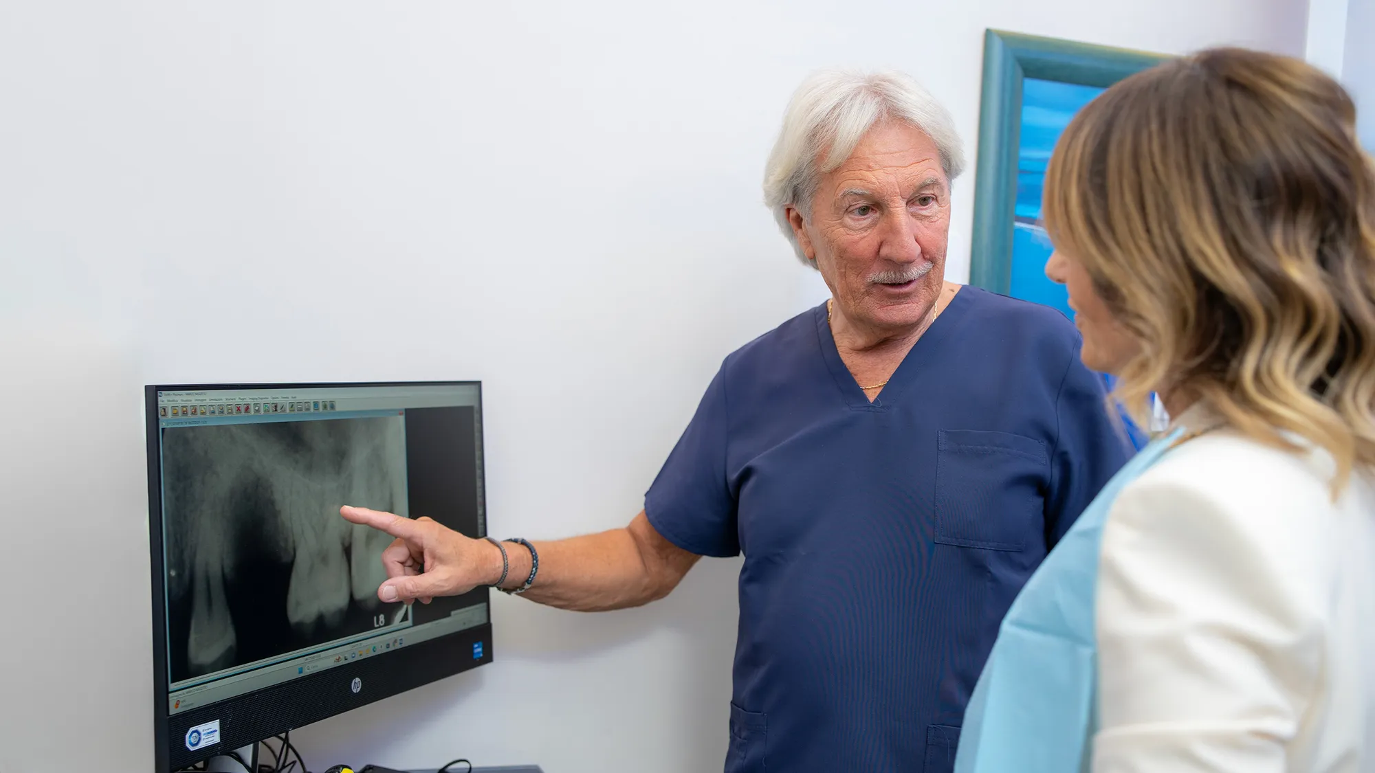 Dentista con camice blu scuro indica una radiografia dentale endorale visualizzata su un monitor, spiegando la diagnosi a una paziente. L'ambiente è uno studio odontoiatrico con pareti bianche e dettagli azzurri.