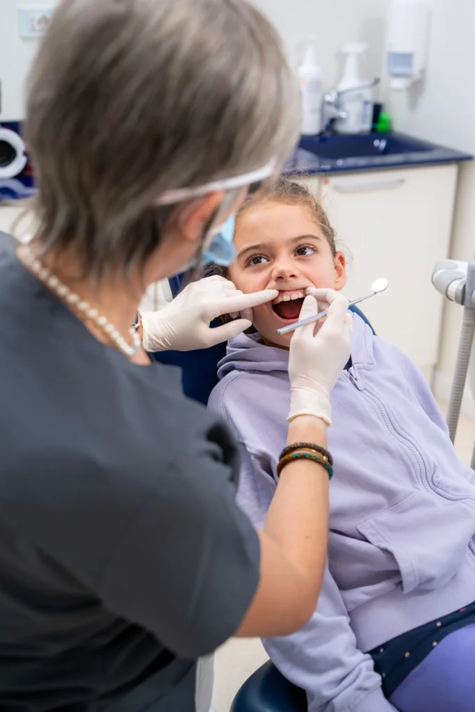 Dentista con guanti bianchi, mascherina e occhiali protettivi visita una bambina con felpa lilla seduta sulla poltrona odontoiatrica. La dottoressa utilizza uno specchietto dentale mentre con l'altra mano allarga delicatamente il labbro superiore per esaminare i denti della piccola paziente, che tiene la bocca aperta con espressione collaborativa.