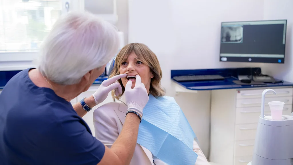 Dentista con camice blu scuro e guanti bianchi esamina la bocca di una paziente seduta sulla poltrona con bavaglio azzurro. Sullo sfondo, un monitor mostra una radiografia dentale e sono visibili le attrezzature dello studio odontoiatrico Dentas.