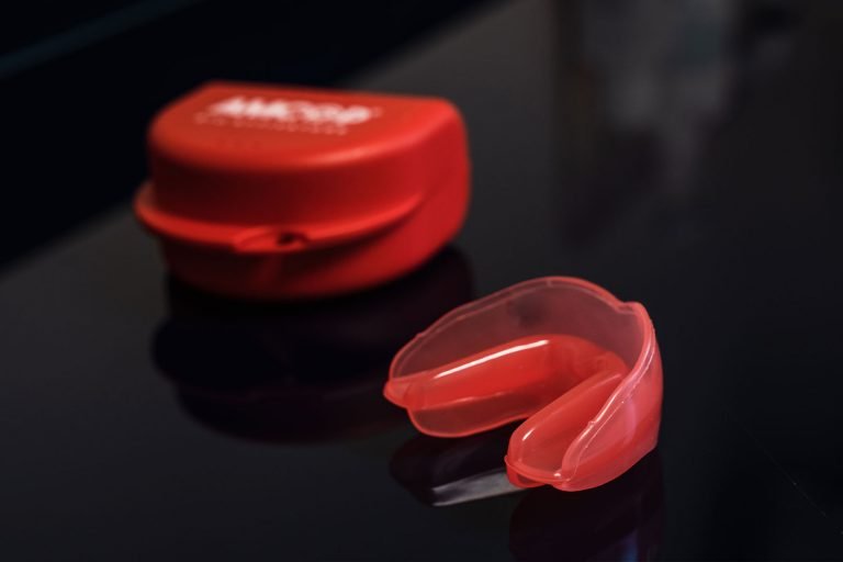 Dispositivo ortodontico miofunzionale in silicone rosso trasparente appoggiato su una superficie scura riflettente, accanto alla sua custodia rossa. Il bite è utilizzato per la valutazione e la correzione di abitudini viziate come la respirazione orale o la deglutizione atipica nei bambini.