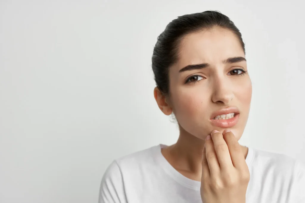 Giovane donna con espressione sofferente si tocca la guancia e il mento con la mano, mostrando segni di dolore dentale. Indossa una maglietta bianca su sfondo chiaro.