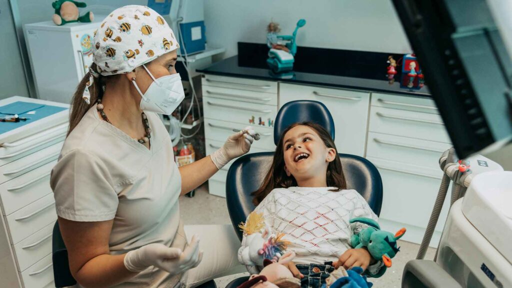 Bambina con capelli castani ride sulla poltrona odontoiatrica blu, stringendo dei peluche tra le braccia, mentre la dentista pediatrica con camice bianco, cuffia con disegni di apine e mascherina tiene in mano uno specchietto dentale. L'ambiente è accogliente, con giocattoli e pupazzi colorati sugli scaffali dello studio.