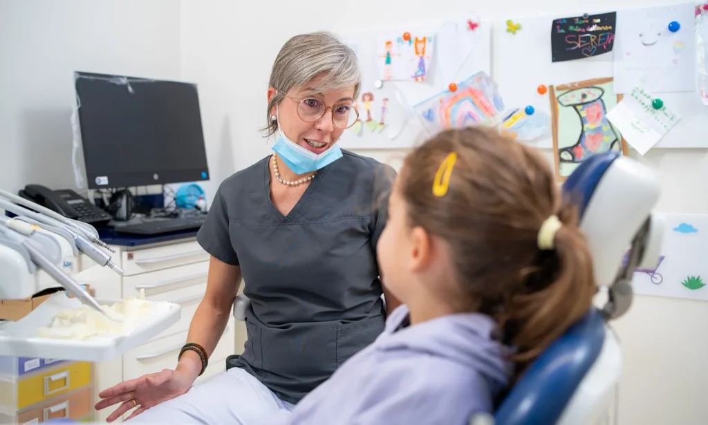 Dentista pediatrica con camice grigio, occhiali, collana di perle e mascherina abbassata parla in modo rassicurante con una bambina seduta sulla poltrona odontoiatrica. Sullo sfondo, una parete decorata con disegni colorati dei piccoli pazienti e un monitor dello studio.
