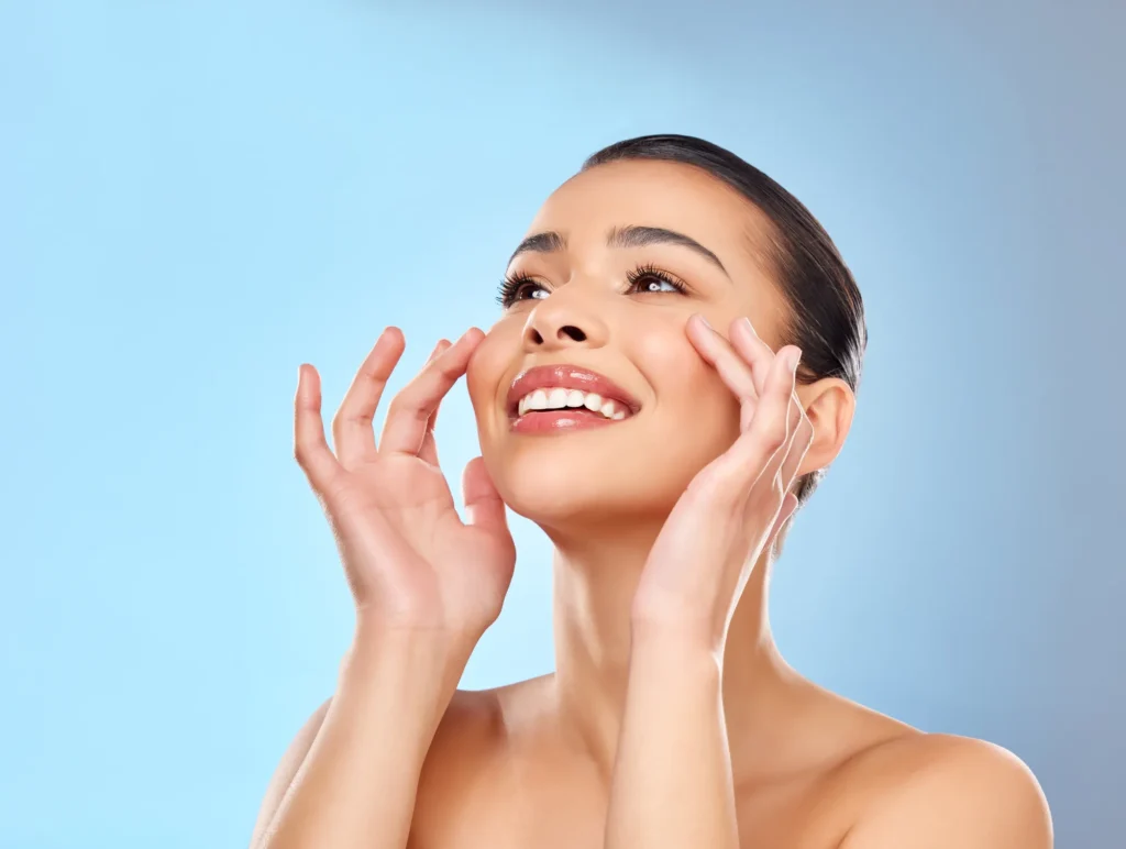 Giovane donna sorridente con pelle luminosa e curata si tocca delicatamente il viso con entrambe le mani, guardando verso l'alto. Lo sfondo è azzurro chiaro e l'immagine comunica benessere e risultati di medicina estetica.