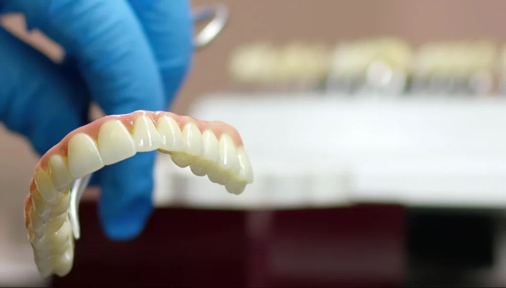Mano con guanto blu tiene una protesi dentale completa ad arcata superiore con denti bianchi e gengiva in resina rosa, all'interno di un laboratorio odontotecnico. Lo sfondo è sfocato con attrezzature di laboratorio.