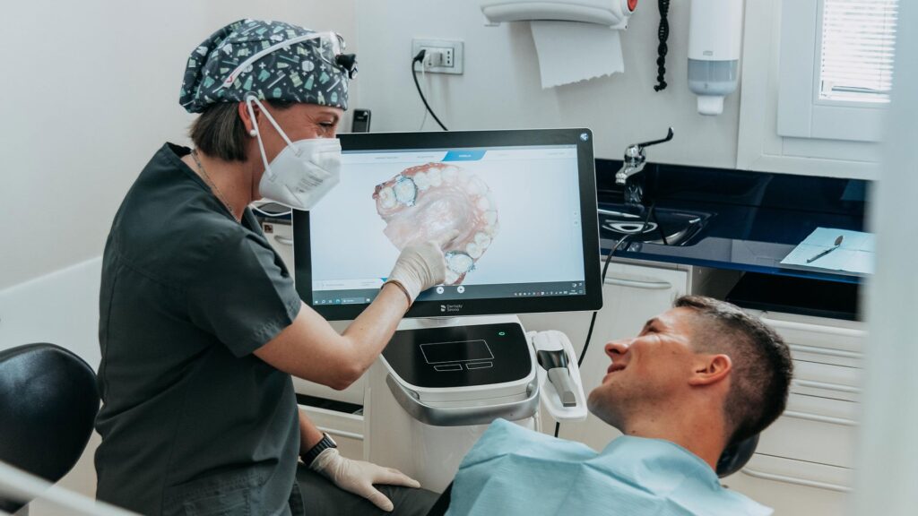 Odontoiatra con camice grigio scuro, cuffia chirurgica e mascherina mostra a un paziente sdraiato sulla poltrona il modello 3D della sua arcata dentale su uno scanner intraorale Dentsply Sirona. Sul monitor è visibile la ricostruzione tridimensionale dell'arcata superiore con dettagli delle superfici occlusali.