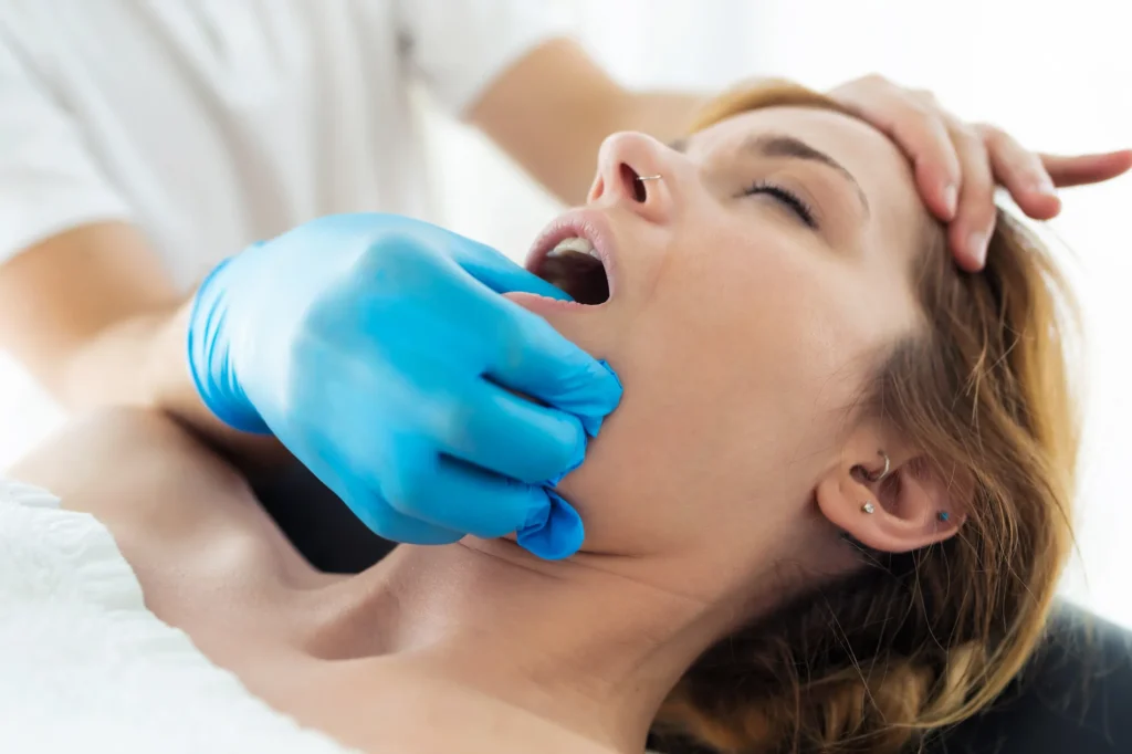 Donna sdraiata con la bocca aperta mentre un professionista con guanti blu esegue un trattamento di massaggio intraorale, con una mano sulla mandibola e l'altra sulla fronte della paziente. La tecnica è utilizzata per il trattamento di disturbi dell'articolazione temporo-mandibolare.