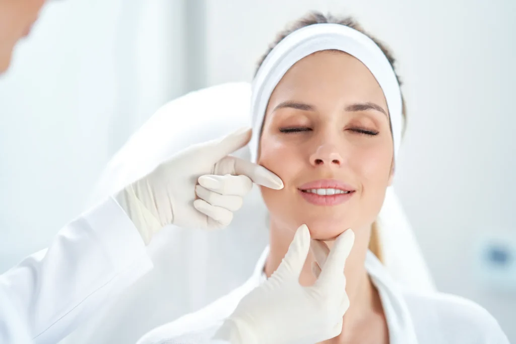 Donna sorridente con gli occhi chiusi e fascia bianca tra i capelli, durante una visita di medicina estetica. Le mani di un professionista con guanti bianchi esaminano delicatamente la pelle del viso, toccando guancia e mento della paziente in un ambiente clinico luminoso.