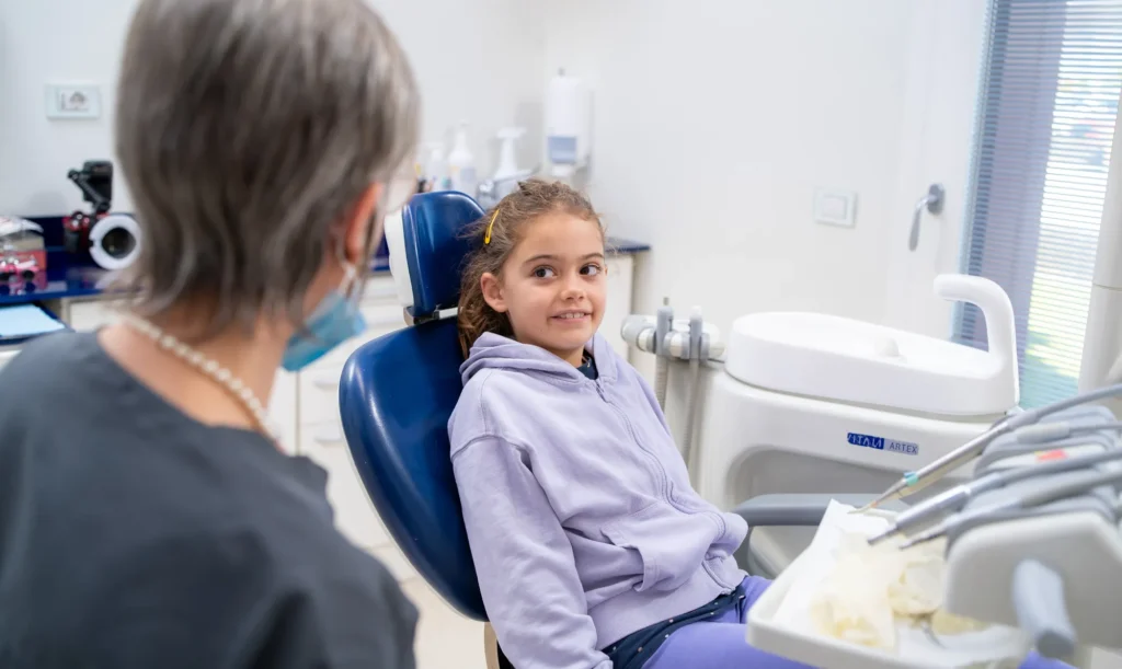 Bambina sorridente con felpa lilla seduta sulla poltrona odontoiatrica blu, guarda con fiducia il dentista di spalle con mascherina abbassata. Sullo sfondo, strumenti odontoiatrici e attrezzature dello studio dentistico.